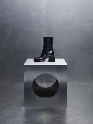 Black leather ankle boot with chunky block heel displayed on a metallic cube table with a circular cutout.