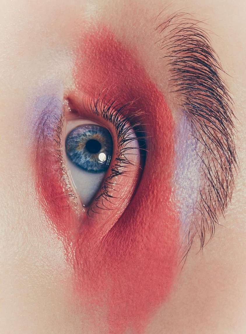 Eye close-up with vivid blue iris, long lashes, and striking red and purple makeup framing the eye area against pale skin.