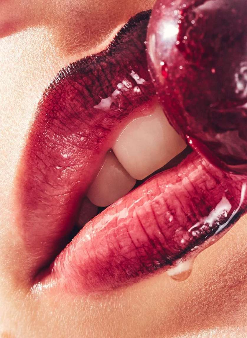 Close-up of a person's mouth with dark red lipstick eating a red lollipop, showcasing the lips, teeth, and skin texture.