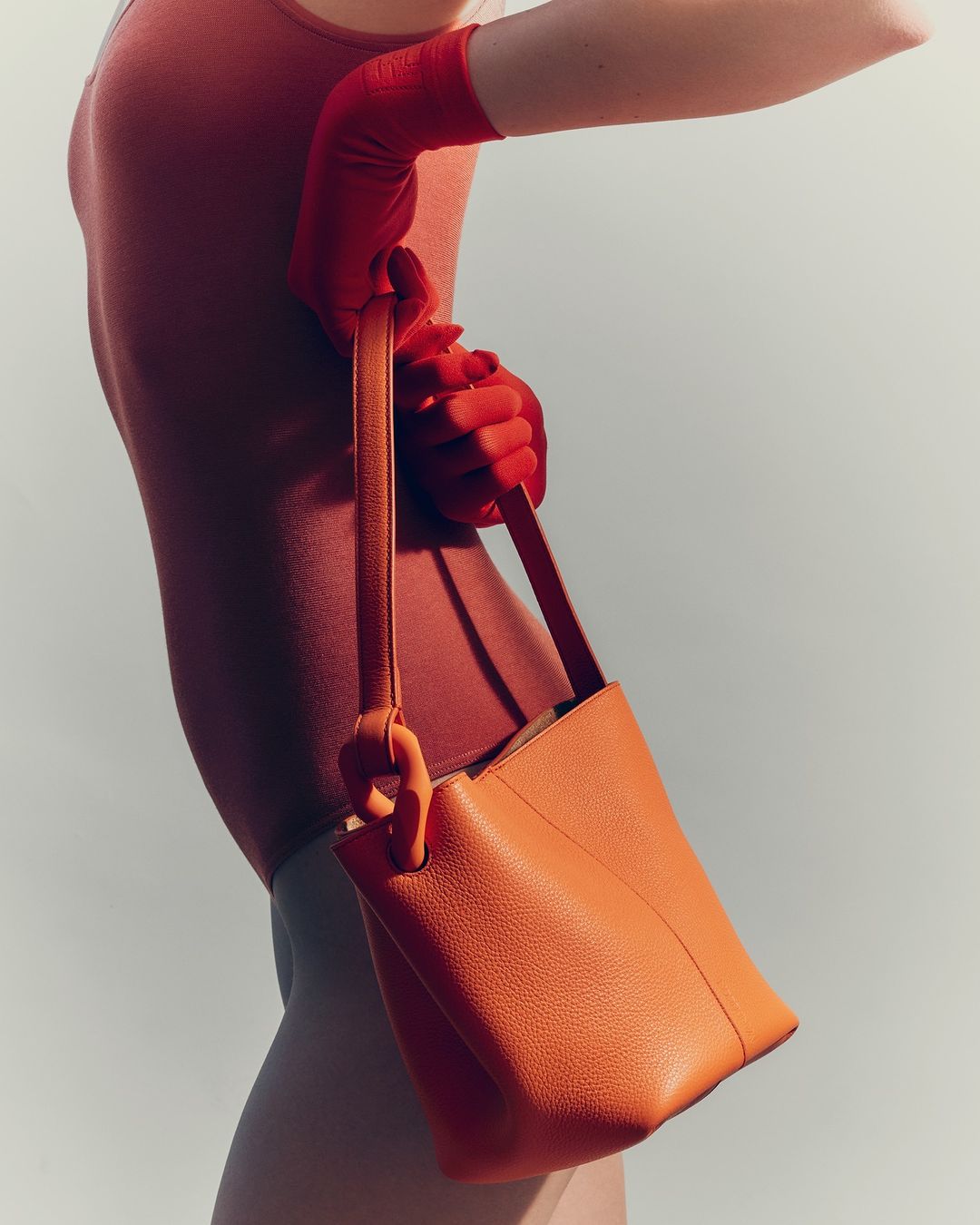Close-up shot of an orange leather bucket bag held by a person wearing a coral bodysuit and red gloves against a light background.