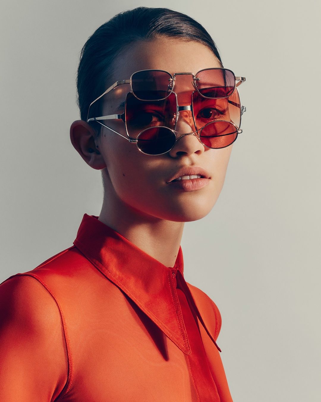 Woman in red blouse wearing layered sunglasses in a fashion editorial shot against a neutral background, medium shot.
