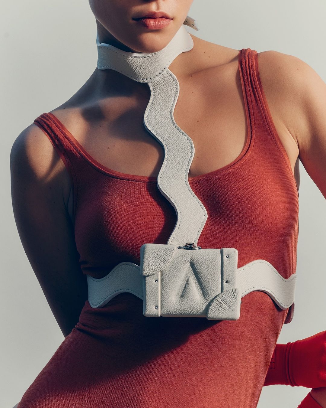 Woman poses wearing rust colored leotard and light-grey leather chest harness with geometric bag, against a white backdrop in a studio.