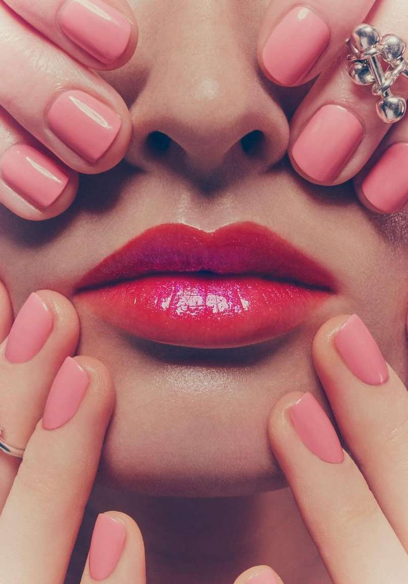 Close-up beauty portrait of a woman's face, focusing on her glossy red lips and manicured pink nails framing her nose and upper lip.