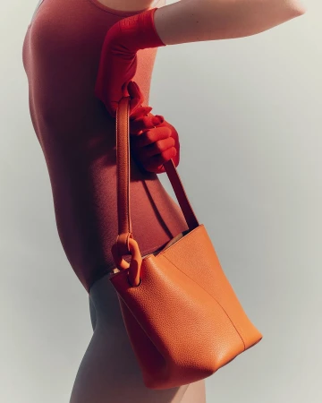 Model wearing red gloves holding an orange leather handbag against a neutral grey background by Romin Favre