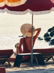 Woman in red swimsuit and wide-brim straw hat sitting under a red fringed beach umbrella by the sea
