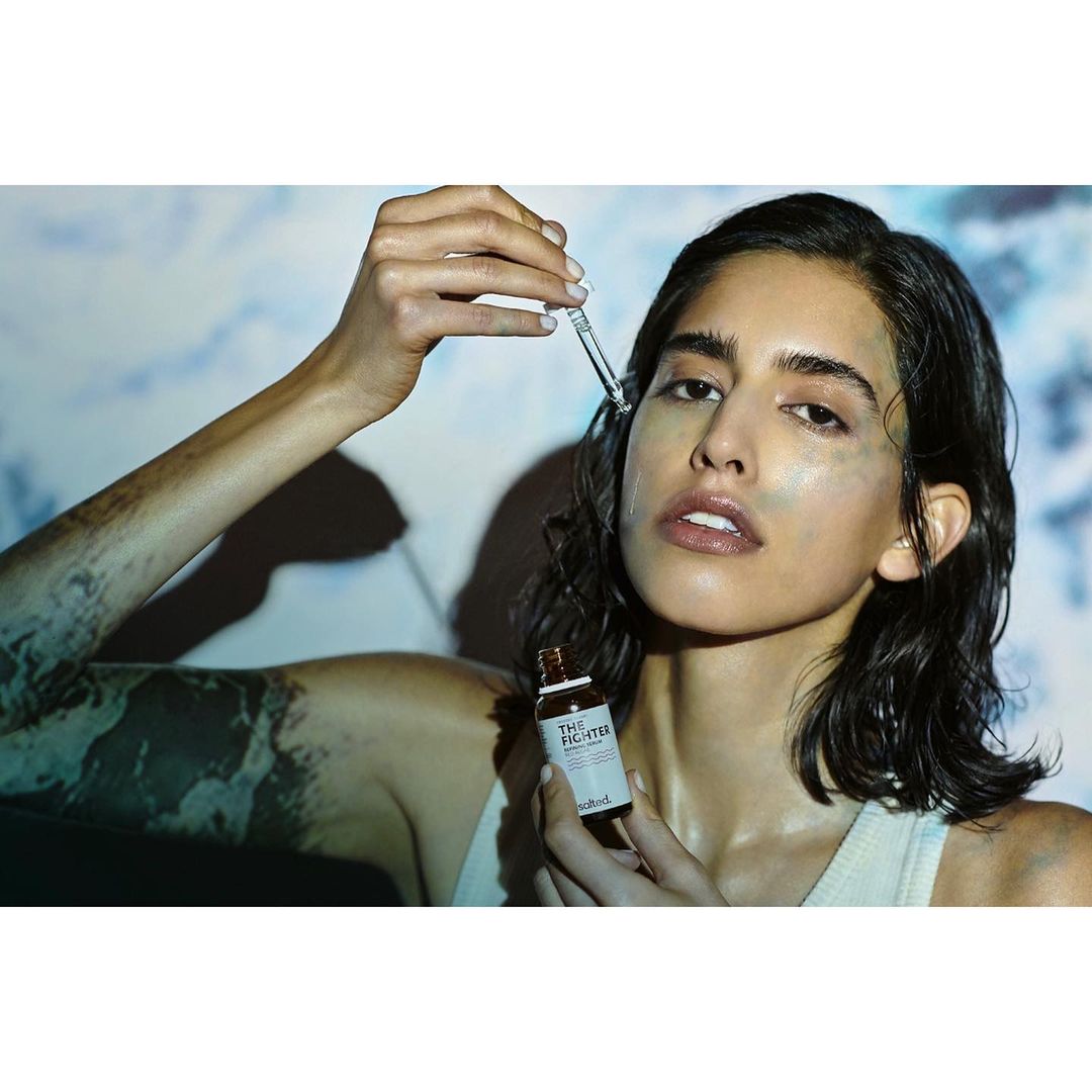 Woman applying serum with dropper on her face, with soft lighting and cosmetic bottle on the foreground against a dreamy backdrop.