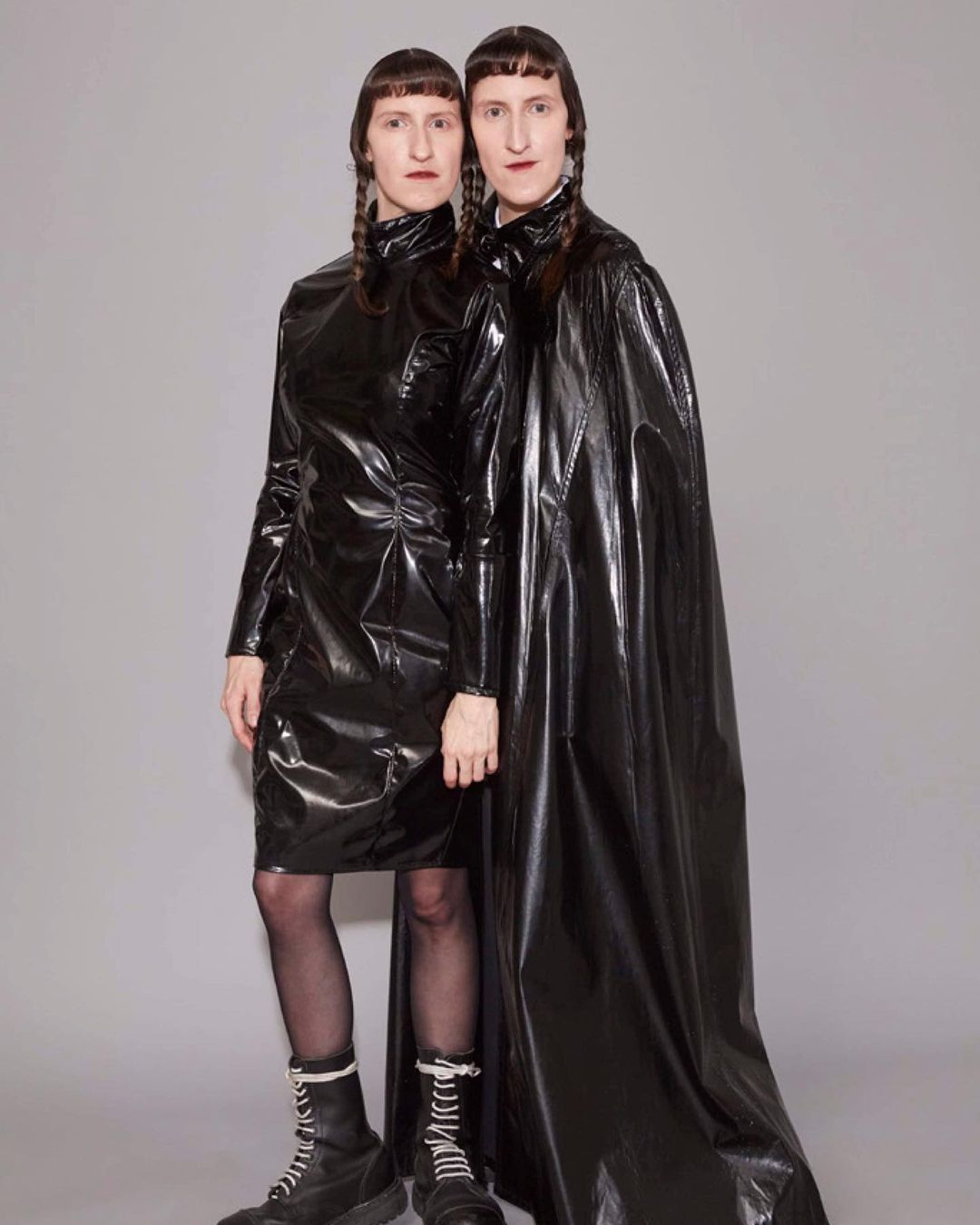 Two adult women in black latex dresses and boots pose in a studio, both with dark braided pigtails and neutral expressions against a grey backdrop.