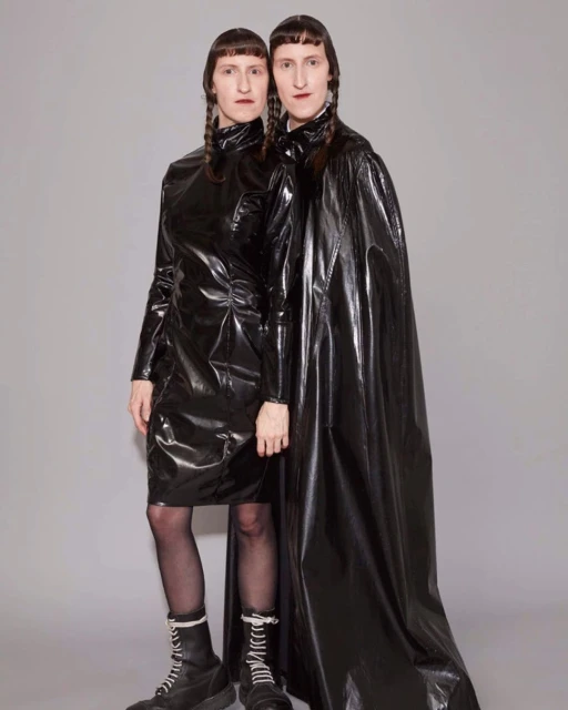 Two adult women in black latex dresses and boots pose in a studio, both with dark braided pigtails and neutral expressions against a grey backdrop.