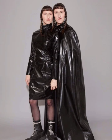 Two adult women in black latex dresses and boots pose in a studio, both with dark braided pigtails and neutral expressions against a grey backdrop.