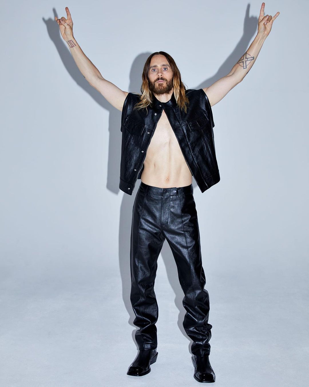 Jared Leto poses in a leather vest, pants, and boots against a white backdrop, giving the 'rock on' hand gesture with raised arms.