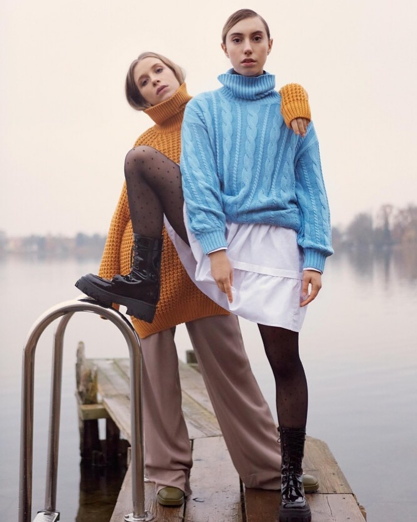 Two women pose on a dock wearing blue and orange turtlenecks, shiny boots, and hosiery; one is standing, the other is leaning on a rail.