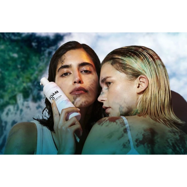 Two adult women pose indoors in front of a light blue and green background, one holding a white foam cleanser product.