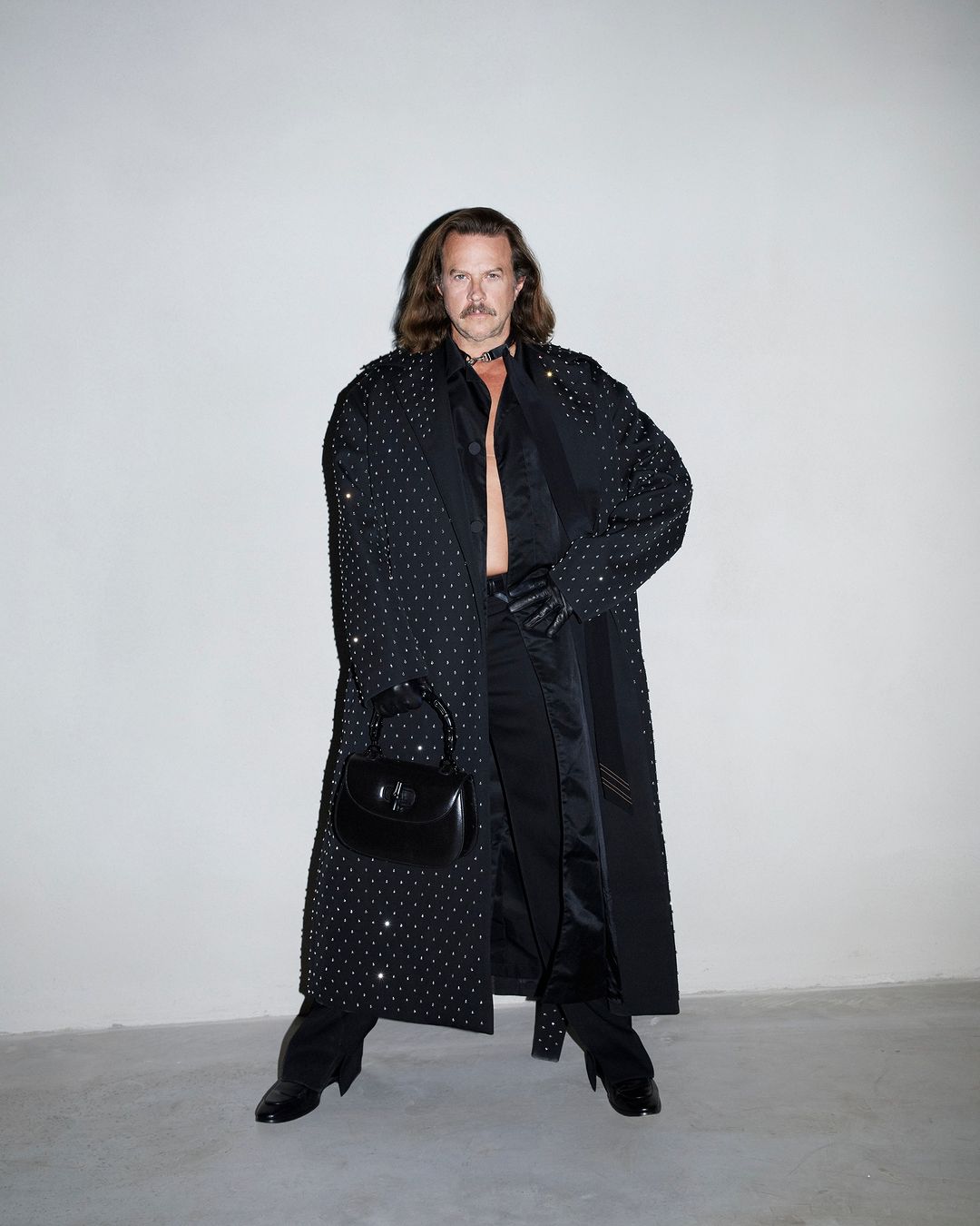 Man with long hair and mustache wearing a crystal-embellished black coat and holding a black handbag against a grey seamless background