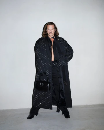 Man with long hair and mustache wearing a crystal-embellished black coat and holding a black handbag against a grey seamless background