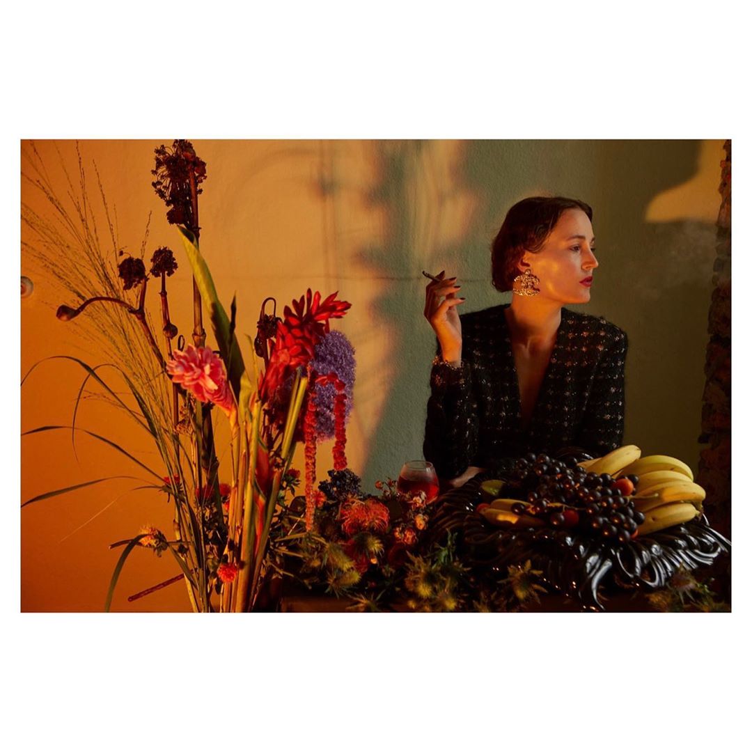Elegant woman smoking in dimly lit interior with lush floral arrangement and fruit, wearing a black tweed jacket and statement earrings.