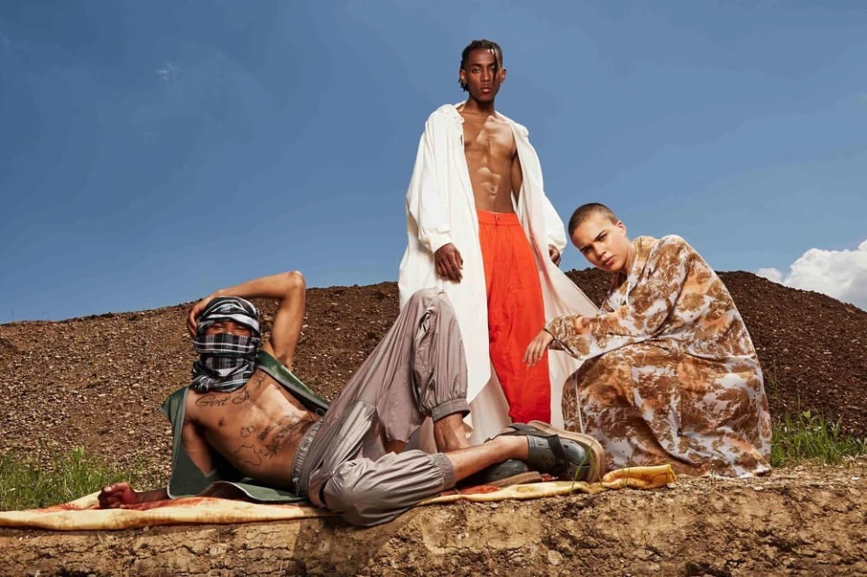 Three fashion models pose outdoors on a dirt mound against a blue sky backdrop, showcasing avant-garde clothing and unique styles.
