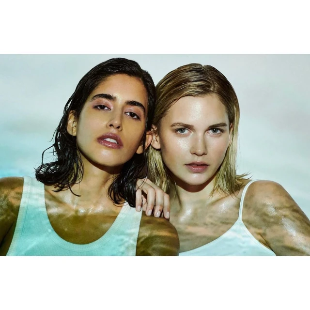 Two young women pose indoors in a close-up beauty shot with wet hair and skin, wearing white tank tops against a dreamy light blue background.