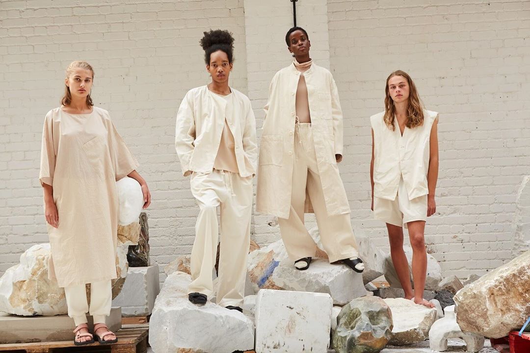 Four fashion models wear neutral-toned clothing while posing on and around stone blocks in a brightly lit indoor setting with white walls.