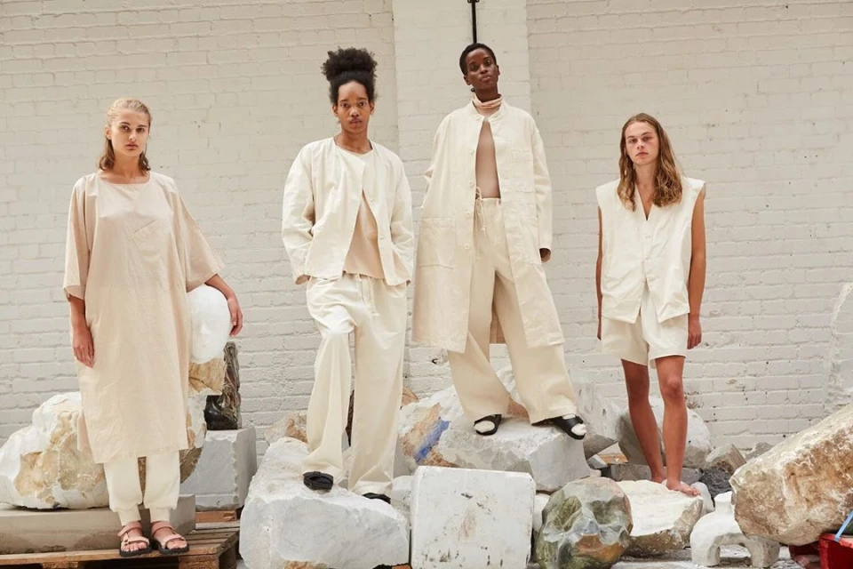 Four fashion models wear neutral-toned clothing while posing on and around stone blocks in a brightly lit indoor setting with white walls.