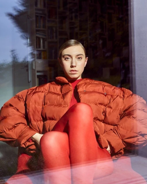 Woman in orange puffer jacket and red leggings sitting indoors with a bookshelf visible through a window, looking at the camera.