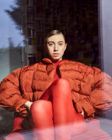 Woman in orange puffer jacket and red leggings sitting indoors with a bookshelf visible through a window, looking at the camera.