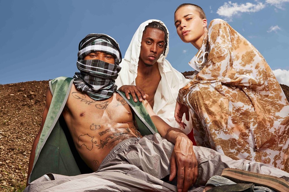 Three models pose outdoors. The model on the left wears a green vest and a head covering, the center model a white hoodie, and the right model a patterned top.