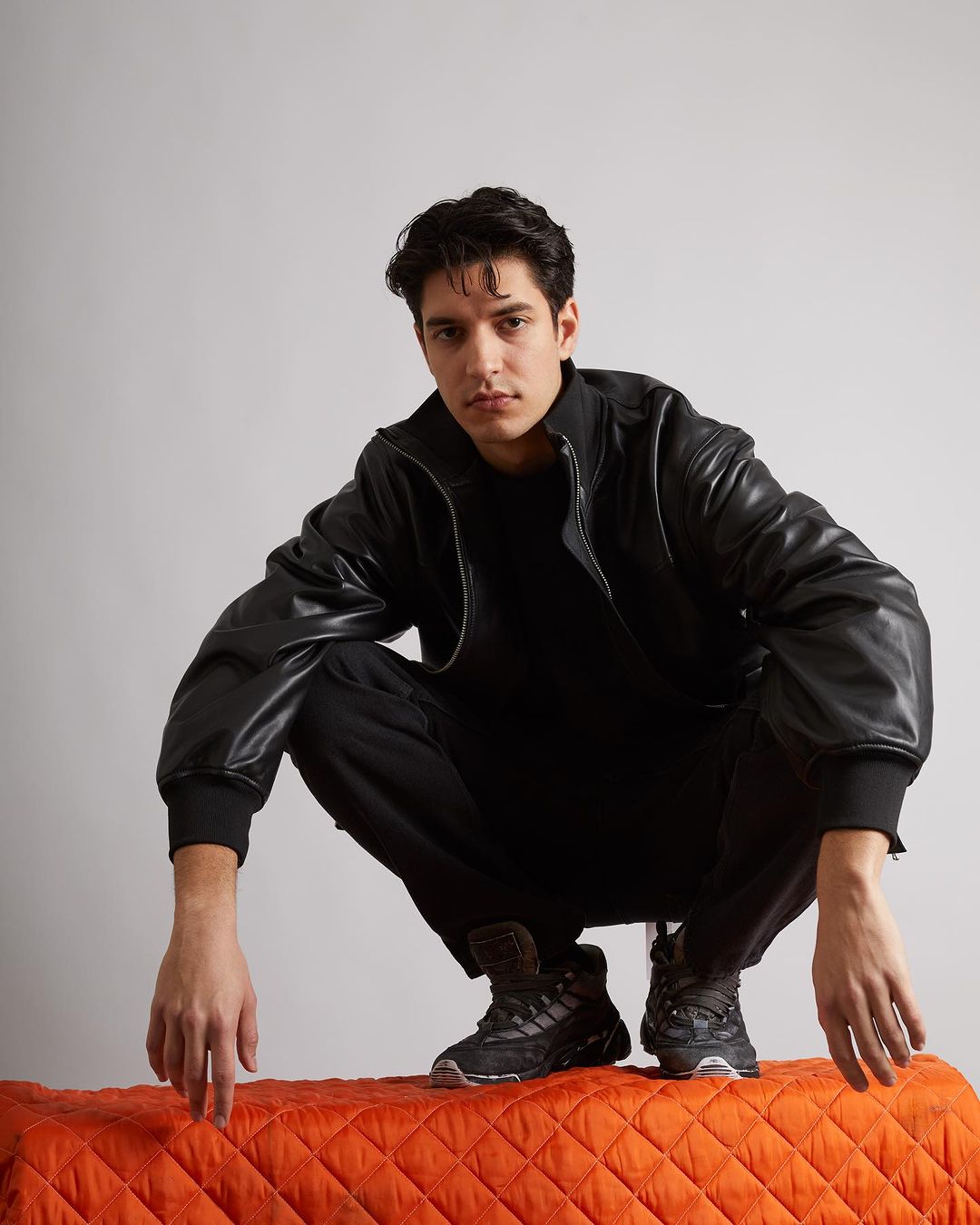 A stylish male model in a black leather jacket poses in a studio setting. The image showcases fashion and contemporary style.
