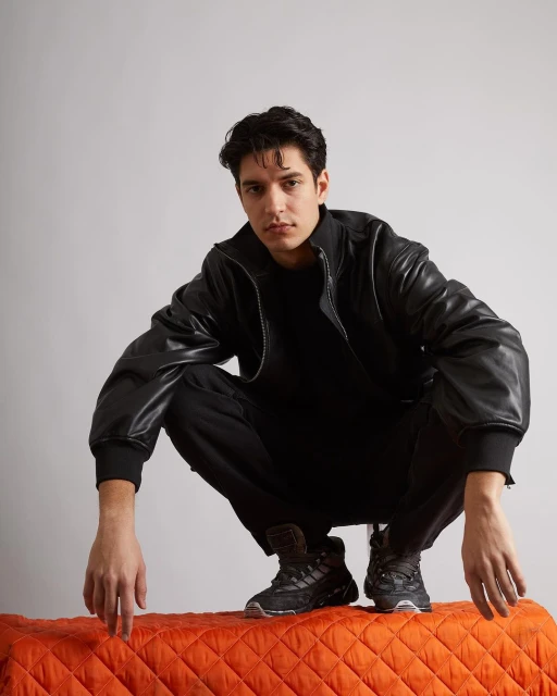 A stylish male model in a black leather jacket poses in a studio setting. The image showcases fashion and contemporary style.