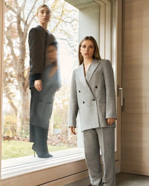 Two women in suits pose indoors with a window and outdoor background. One woman is a reflection. Fashion editorial shot.