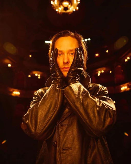 Man in a dark coat poses indoors with hands framing his face. The background is dark with light fixtures visible in the distance.