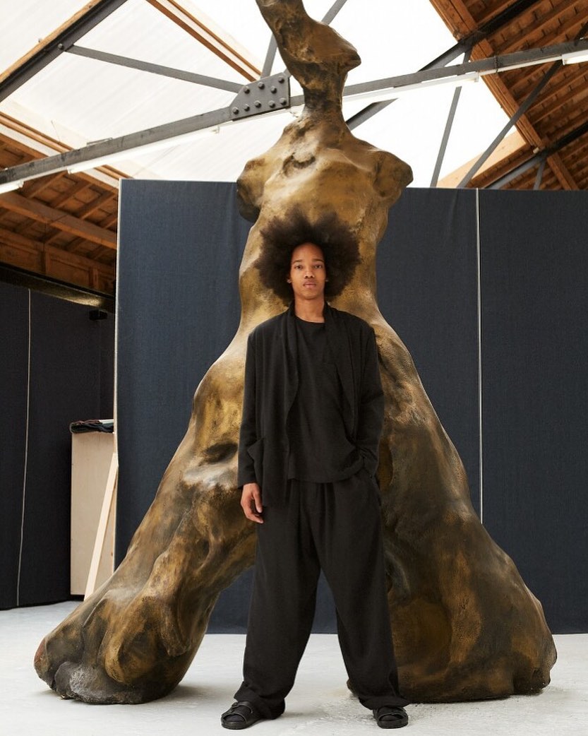 Studio shot of a young adult posing next to a large sculpture in an indoor setting with natural light and a dark background.