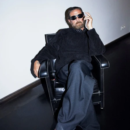 Man with beard wearing sunglasses and black sweater seated in a black armchair against a plain wall