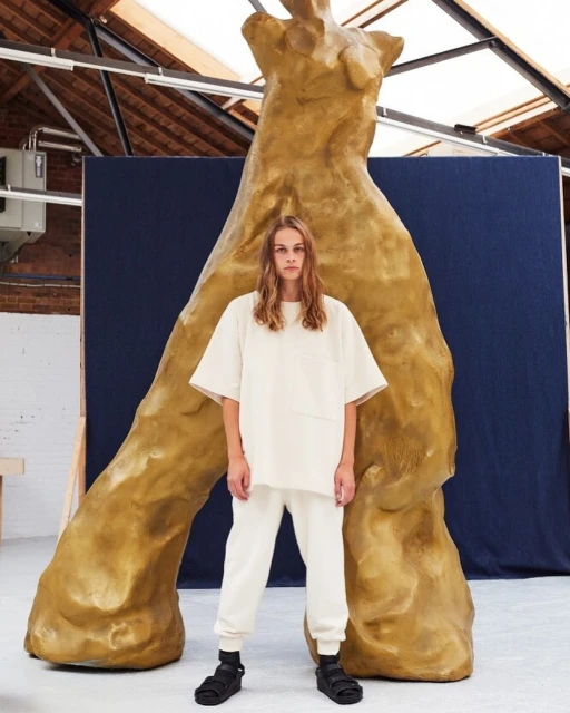 A slender adult male stands in front of a large tan sculpture wearing a matching short-sleeve shirt and sweatpants in a bright indoor space.