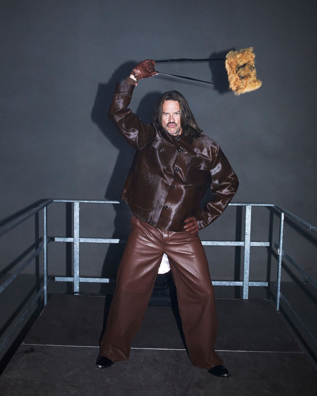 Adult male in a brown jacket and pants posing on a metal platform, holding a fuzzy object overhead, set against a dark gray backdrop.