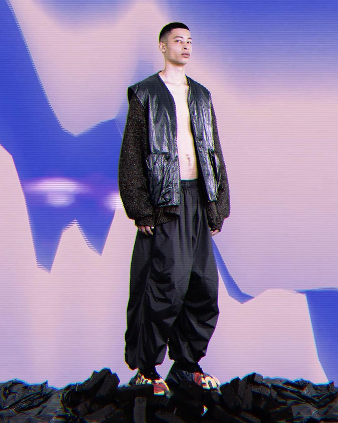A male model wearing a dark leather vest over textured knit sleeves and loose black trousers standing on a dark rocky ground against a colorful abstract gradient background.