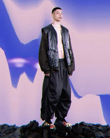 A male model wearing a dark leather vest over textured knit sleeves and loose black trousers standing on a dark rocky ground against a colorful abstract gradient background.