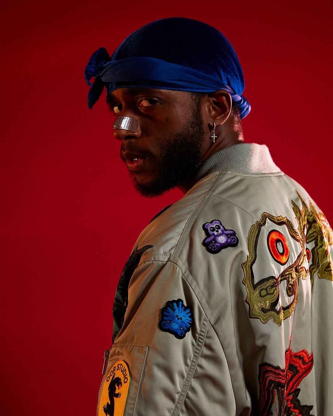 Man in blue headscarf and patched jacket posing against red background. Portrait of male with bandages on nose looking over shoulder.
