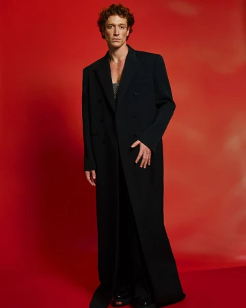 Man with curly brown hair wearing a long black coat standing against a solid red studio background.