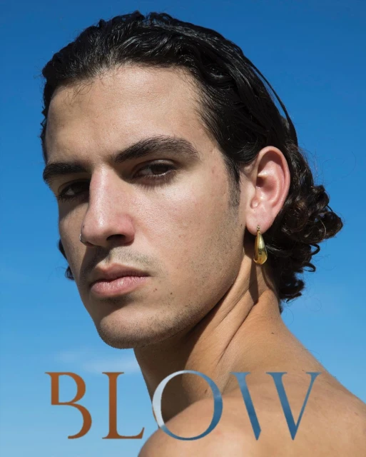 Portrait of a light-skinned male with dark curly hair, wearing an earring, against a clear blue sky. The text 'BLOW' is at the bottom.