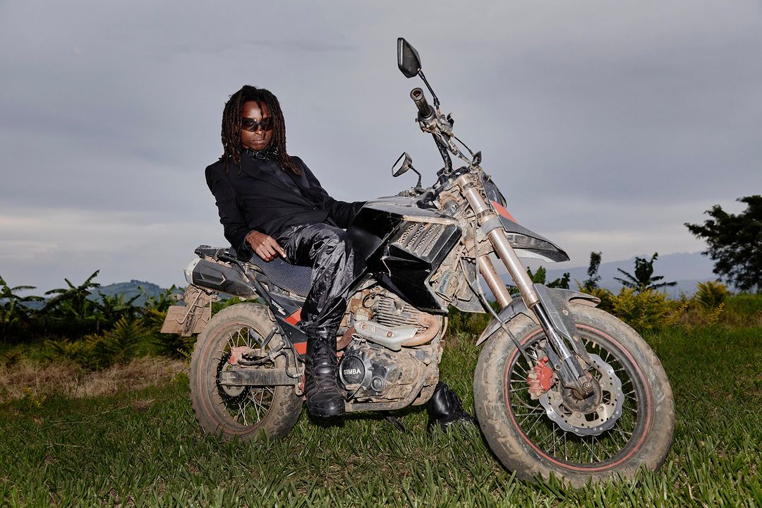 Adult male with dreadlocks in a dark suit is posing while sitting on a black and silver dirt bike in a grassy, rural landscape.