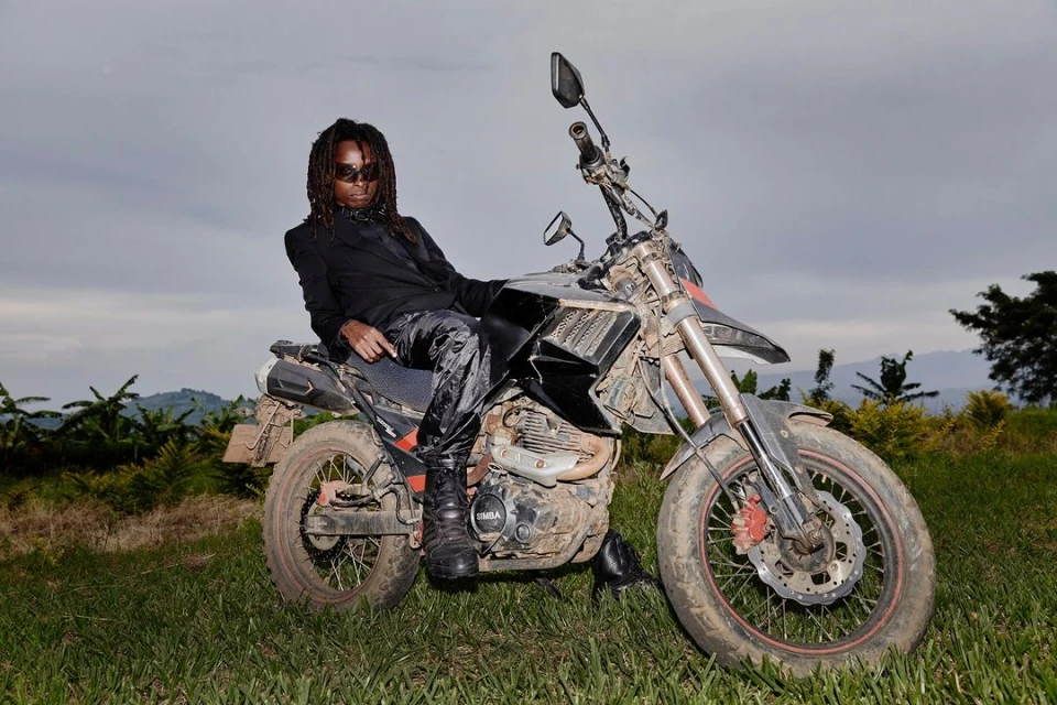 Adult male with dreadlocks in a dark suit is posing while sitting on a black and silver dirt bike in a grassy, rural landscape.