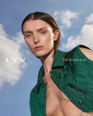 Fashion portrait of a woman with neutral expression, wearing a green vest, against a blue sky with clouds, fashion, beauty, outdoor.