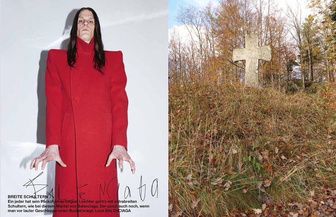 Model in a red coat with exaggerated shoulders in a studio next to an outdoor scene with a stone cross in a grassy field.