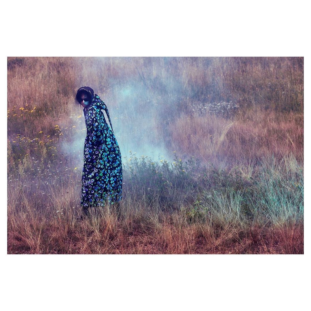 Woman in a floral dress stands in a misty field of tall grass, flowers, and weeds, with a muted color palette and soft lighting.