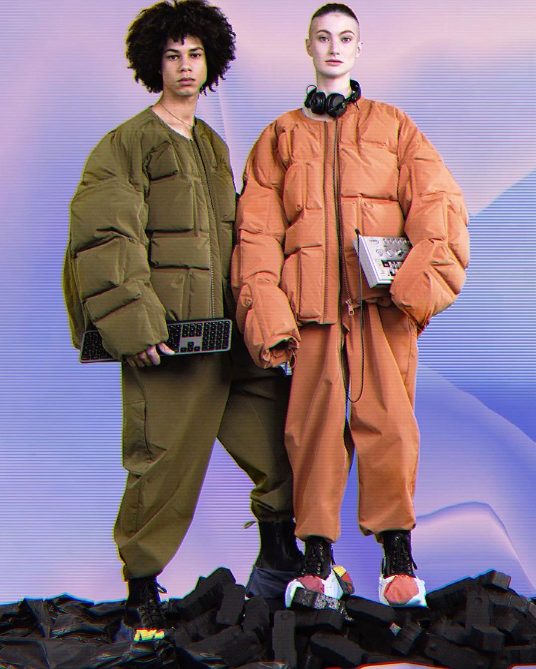 Two fashion models wear oversized olive green and orange puffer jackets, standing on a pile of black brick debris in a photo studio.