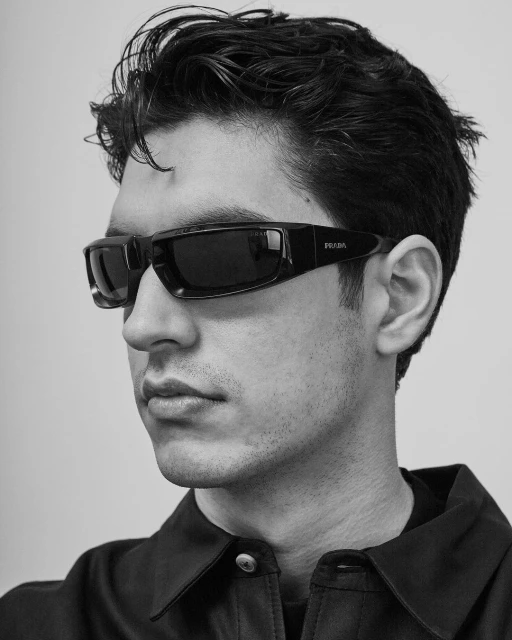 Monochrome portrait of a man wearing Prada sunglasses and a dark collared shirt against a plain backdrop. Head and shoulders shot.