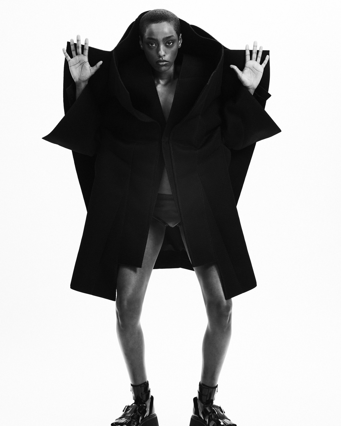 Fashion shot of a Black female adult wearing a black oversized coat and black boots, standing in a photo studio against a white backdrop.