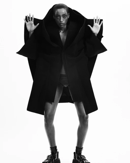 Fashion shot of a Black female adult wearing a black oversized coat and black boots, standing in a photo studio against a white backdrop.