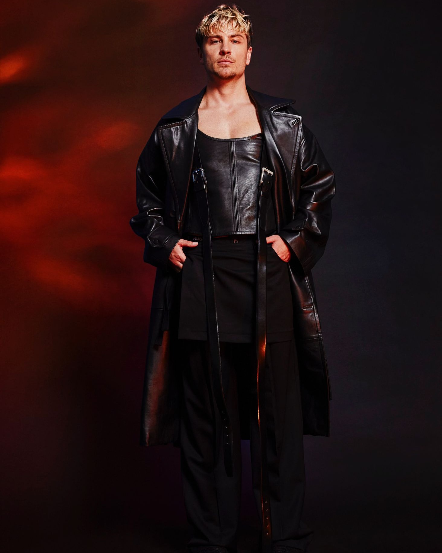Adult male fashion model in black leather top and long coat poses in a studio with a red and black gradient background.