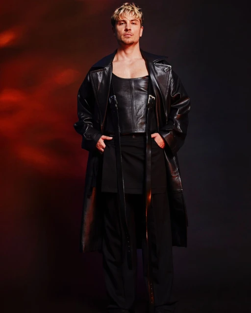 Adult male fashion model in black leather top and long coat poses in a studio with a red and black gradient background.